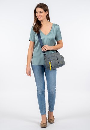 Borsa a tracolla in tessuto grigio con due tasche frontali, zip gialla, e tracolla regolabile, mostrata su una persona in abbigliamento casual.