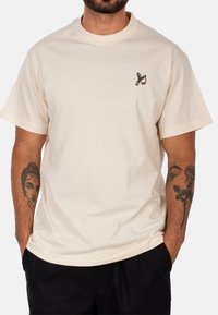 T-shirt a maniche corte di colore crema realizzato in cotone, caratterizzato da un piccolo design ricamato sul petto. Vestibilità ampia con scollo rotondo.