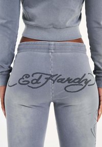 Grijze katoenen joggers hebben een groot zwart 'Ed Hardy' logo aan de achterkant. De tailleband is geribbeld en de stof heeft een soepele textuur.