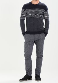 Marinblått stickat tröja med vita geometriska mönster, i kombination med grå texturerade byxor. Svarta skor fulländar outfiten.