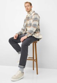 Kariertes Flanellhemd in Creme, Grau und Beige; graue Hosen mit aufgeschlagenen Säumen; cremefarbene Sneaker mit beigen Akzenten; sitzend auf einem Holzschemel.