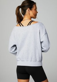 Sweat-shirt oversize gris clair avec un grand col et des épaules tombantes. Porté par-dessus des bretelles sportives noires. Un short noir ajusté en dessous.