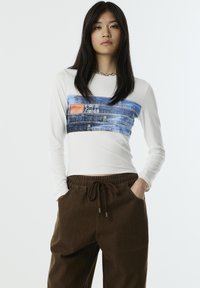 Witte langslijp top met denim grafisch ontwerp en kleine oranje patch; gecombineerd met donkerbruine losse broek met trekkoord in de taille.
