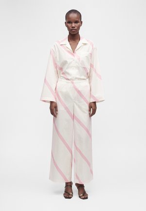 Personne portant une tenue blanche ample avec de larges rayures diagonales roses, de longues manches larges, un pantalon à taille élastique et des sandales noires à bouts ouverts.