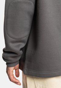 Sudadera gris con una textura suave, puños acanalados, parches en los codos y un ajuste relajado, combinada con pantalones de color claro.