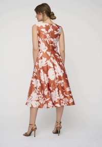 Ein ärmelloses Kleid in knielanger Länge in Rostfarben mit einem weißen Blumenmuster. Das Kleid hat ein tailliertes Oberteil und einen ausgestellten Rock, sowie einen verdeckten Rückenreißverschluss.