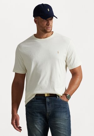 Crème katoenen t-shirt met ronde hals en korte mouwen, voorzien van een klein geborduurd logo. In combinatie met donkere spijkerbroeken en een marineblauwe pet.