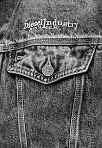 Pilka džinsinė striukė su aštriu krūtinės kišenių, sidabrinių sagų detalėmis ir „Diesel Industry“ siuvinėtu logotipu viršutiniame kairiajame kampe.