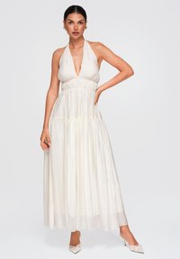 Femme en robe longue blanche sans manches à col halter avec taille froncée, debout, une main sur la hanche et portant des talons blancs à bouts pointus.