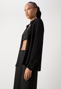 Black long-sleeve shirt made of textured fabric, featuring a button-up design and side slits. Worn over a fitted black crop top and flowing pants.
