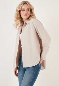 Hele beež pluus, millel on relaxed fit, pikad varrukad ja kaardus allserv. Kombineeritud sobivate siniste teksadega. Sile tekstuur.