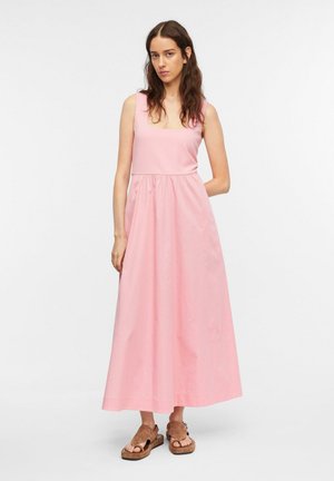 Junge Frau mit langen braunen Haaren, die ein ärmelloses pinkes Maxikleid und braune flache Sandalen trägt, steht vor einem weißen Hintergrund.