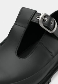 Μαύρο παπούτσι με στρογγυλή μύτη, σχέδιο με κοψίματα, ασημένιο κούμπωμα και λεία, σαν καουτσούκ, υφή. Ραφές κατά μήκος των άκρων.