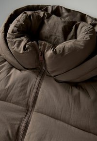 Veste rembourrée marron avec un col haut et froncé et une fermeture éclair sur le devant. Elle présente un tissu lisse avec des sections texturées et de subtils détails de couture.