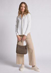 Bolso de cuero marrón suave con forma estructurada, que cuenta con un asa corta y una sutil marca. El modelo lleva una blusa blanca y pantalones beige.