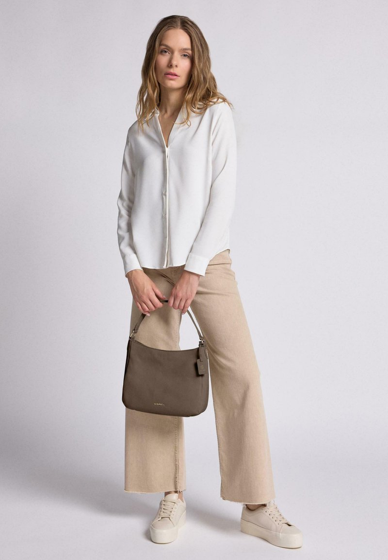 Bolso de cuero marrón suave con forma estructurada, que cuenta con un asa corta y una sutil marca. El modelo lleva una blusa blanca y pantalones beige.