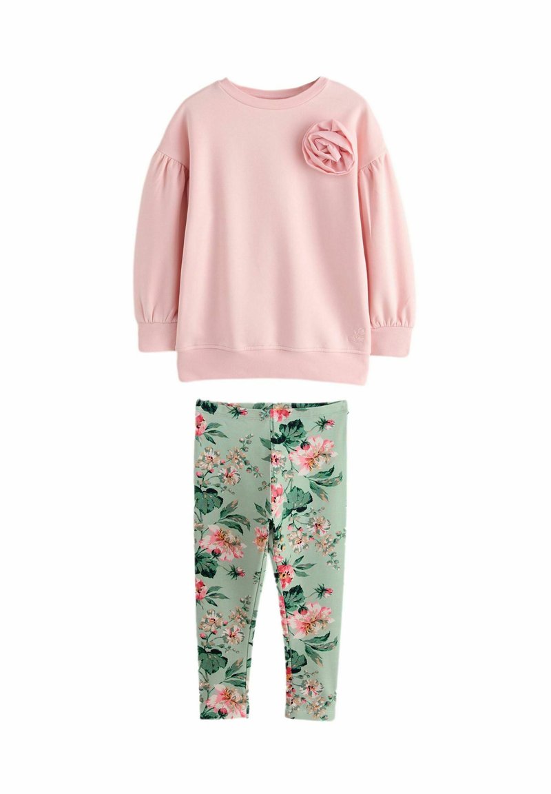Svetlo roza pulover s napihnjenimi rokavi in detajem rožice, v kombinaciji z zelenimi cvetličnimi legginsom, ki prikazujejo roza in bele cvetove.