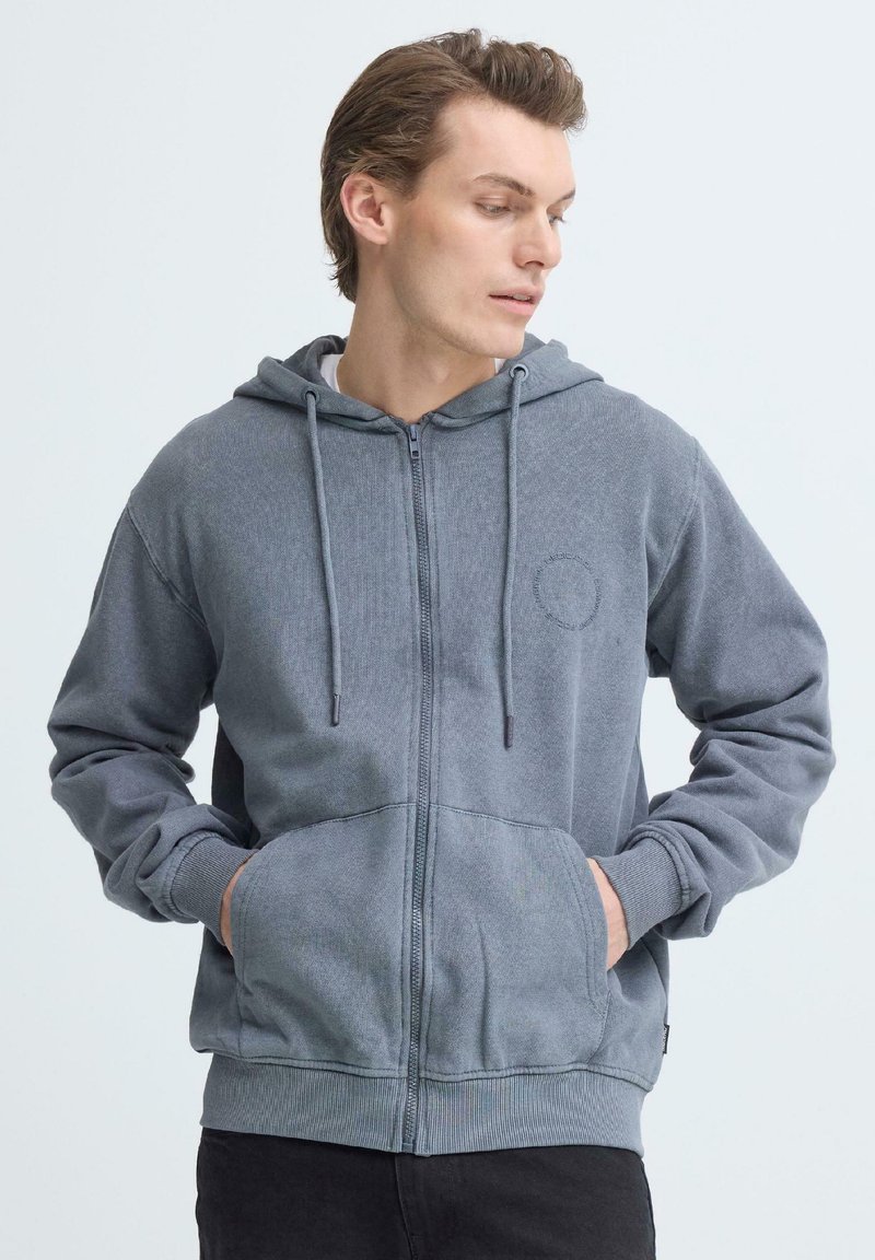 Sweat à capuche gris zippé avec capuche, poches avant et poignets côtelés. Caractéristiques d'un tissu texturé et d'un logo circulaire sur le côté gauche.