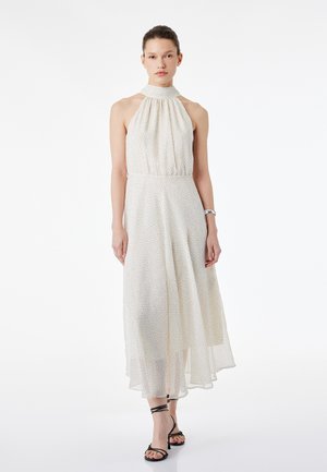 Kobieta ubrana w białą sukienkę midi na szyję bez rękawów w drobne czarne kropki oraz w czarne sandały na wysokim obcasie z paskami, stojąca.