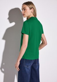 Grön polo shirt med krage, korta ärmar och en mjuk textur, parat med mörka denimbyxor. Skugga synlig på väggen.