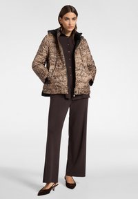 Giacca puffer in stampa leopardata marrone con cappuccio nero rimovibile, zip frontale e tasche, indossata sopra una maglietta scura e pantaloni a gamba larga.
