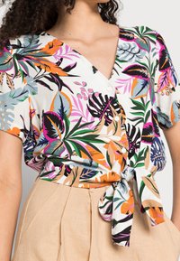 Short-sleeved, white blouse with colorful tropical leaf prints. Features a V-neckline and a tied waist. Fabric appears lightweight and breathable.