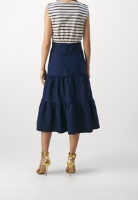 Mujer de pie con la espalda visible, vestida con una blusa sin mangas de rayas, una falda midi en capas de color azul marino y sandalias de tacón alto doradas.