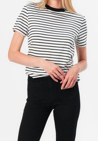 Femme portant un t-shirt rayé horizontal noir et blanc, rentré dans un pantalon noir taille haute, debout les mains près de la taille.