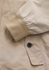 Bézs dzseki sima, fényes külsővel. Az ujjatlan részen világosbarna anyag található. Sötét gombbal és zsebrészletekkel ellátva.