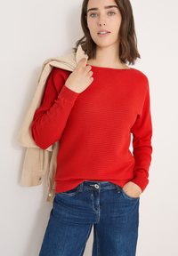 Maglione rosso a costine con maniche lunghe, caratterizzato da un collo a barca, abbinato a jeans blu. Giacca in corduroy crema appoggiata su una spalla.