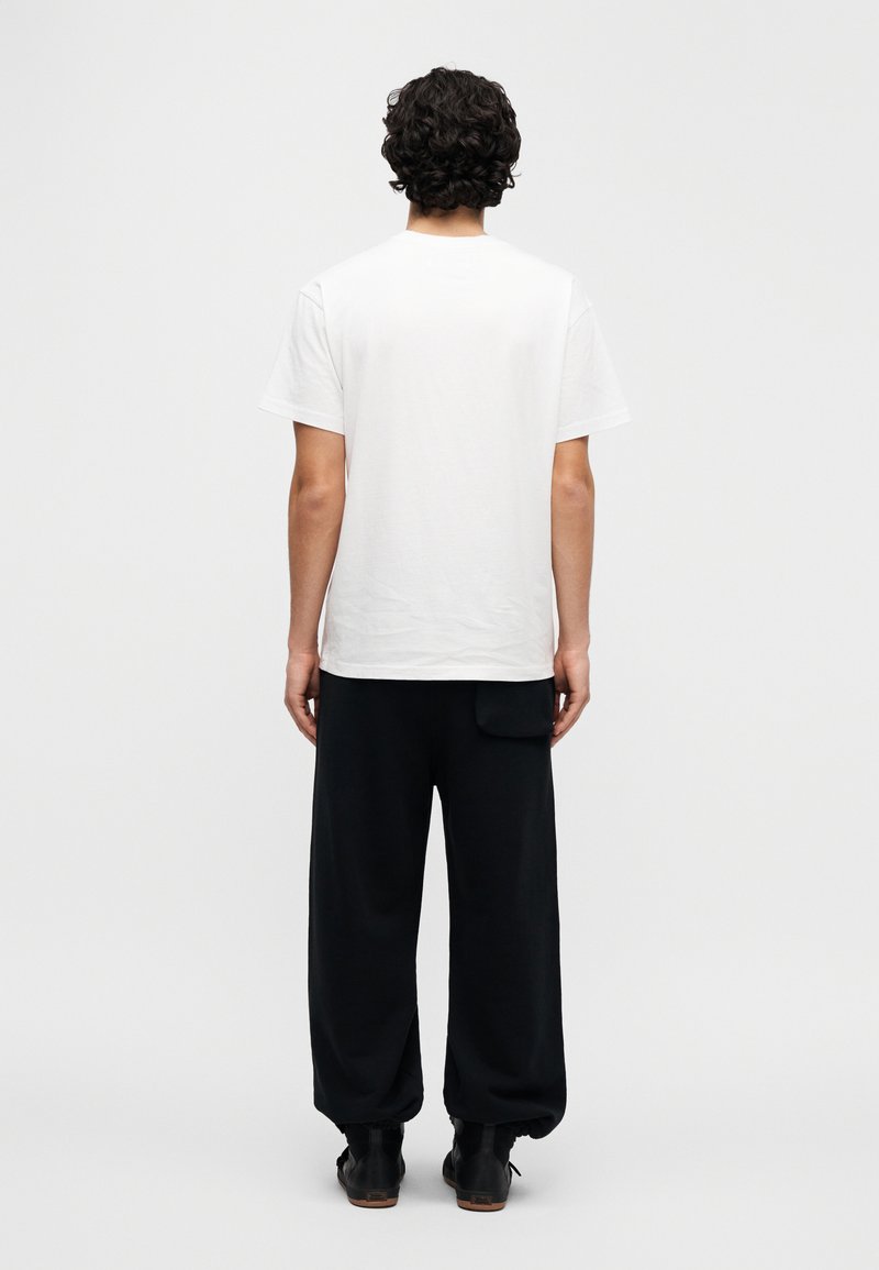 Hvid t-shirt i bomuld med korte ærmer, kombineret med løse sorte bukser. Bukserne har en sidelomme og elastiske manchetter. Sorte sko bæres.