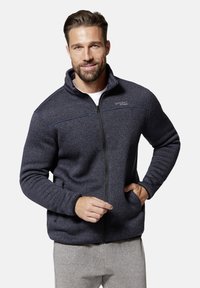 Lächelnder Mann, der eine marineblaue Fleecejacke mit Reißverschluss und hellgraue Hose trägt, mit einer Hand in der Tasche vor einem schlichten, hellen Hintergrund.