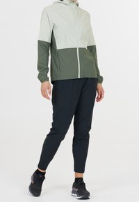 Veste légère bicolore en vert et gris clair, avec fermeture éclair à l'avant et capuche. Associée à un pantalon noir cintré et des chaussures de sport noires.