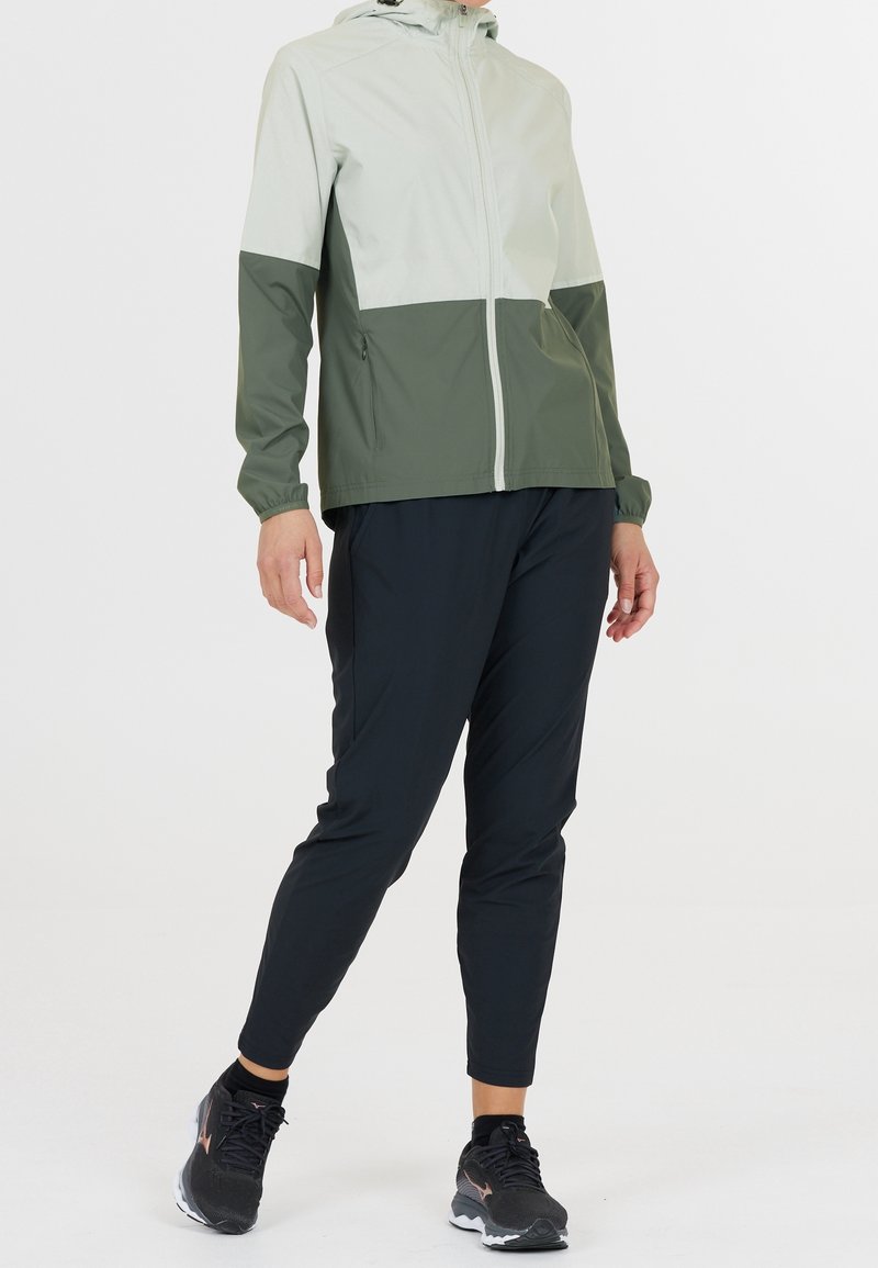 Giacca leggera bicolore verde e grigio chiaro, con zip frontale e cappuccio. Abbinata a pantaloni neri a sigaretta e scarpe da ginnastica nere.