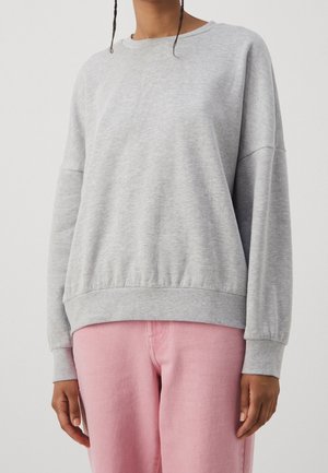 Personne portant un sweat-shirt gris clair à manches longues et un pantalon rose, visible du cou jusqu'aux hanches sur un fond clair uni.