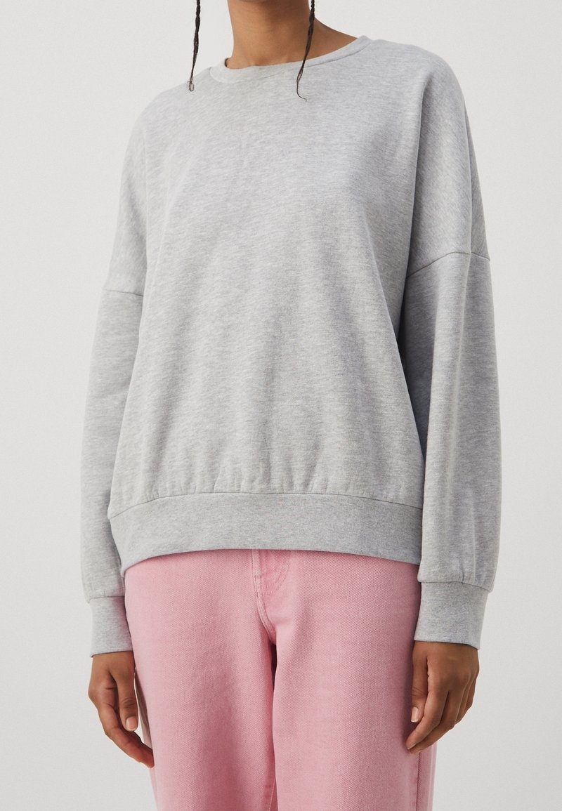 Personne portant un sweat-shirt gris clair à manches longues et un pantalon rose, visible du cou jusqu'aux hanches sur un fond clair uni.