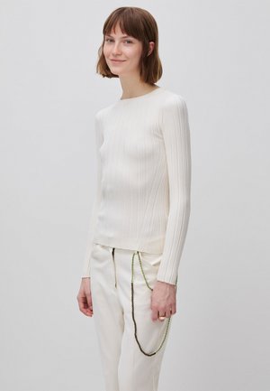 Kobieta o krótkich brązowych włosach, ubrana w biały prążkowany top z długim rękawem i białe spodnie z dwoma ozdobnymi łańcuszkami wiszącymi z kieszeni.