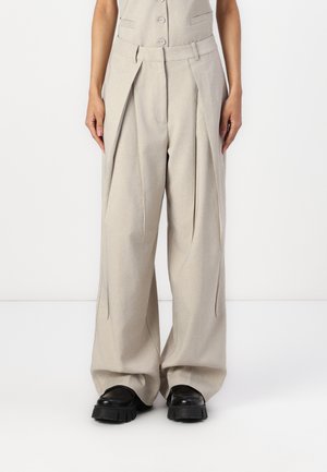 Pantalon beige à jambes larges avec plis, porté avec des chaussures noires à semelles épaisses, montré de la taille aux pieds sur un fond uni.
