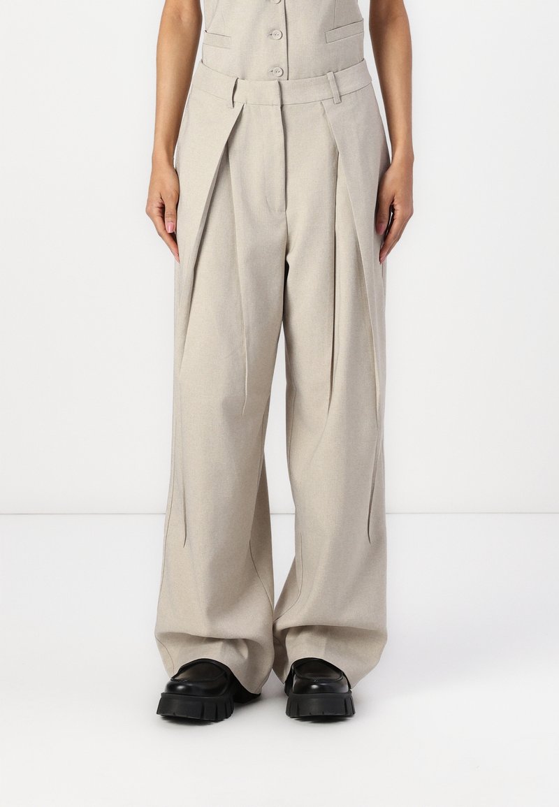Pantalon beige à jambes larges avec plis, porté avec des chaussures noires à semelles épaisses, montré de la taille aux pieds sur un fond uni.