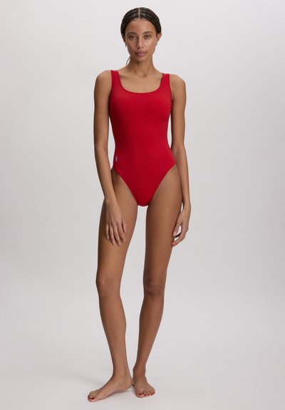 Γυναίκα που στέκεται ξυπόλυτη φορώντας ένα αμάνικο κόκκινο ολόσωμο μαγιό μπροστά σε ένα απλό ανοιχτό φόντο.