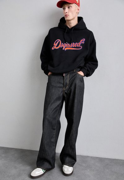 Tânăr îmbrăcat cu un hanorac negru Dsquared2, blugi negri cu picioare largi, adidași albi cu dungi verzi și o șapcă roșie, stând în fața unui zid simplu.