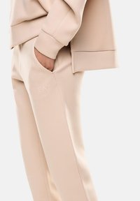 Pantaloni sportivi beige con vestibilità rilassata, dotati di tasche laterali e un logo "EA7" in rilievo sulla coscia, realizzati in un morbido materiale tessile.