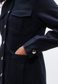 Άτομο που φοράει σκούρο ναυτικό σακάκι με ασημένια κουμπιά, το χέρι του σε μπροστινή τσέπη, δείχνοντας λεπτομερείς πτερύγια τσέπης και κουμπί στο μανίκι.