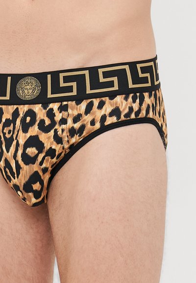 Nahaufnahme einer Person in Leopardenmuster-Unterhosen mit einem schwarzen Bund, der ein goldenes griechisches Schlüsselmotiv und ein Medusa-Logo aufweist.