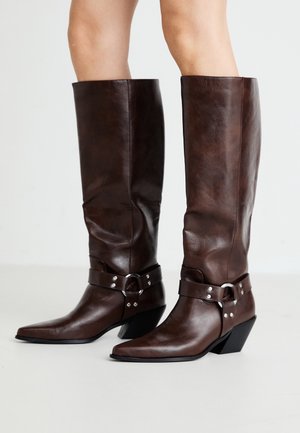 Bottes en cuir brun foncé montantes jusqu'aux genoux avec des bouts pointus, des brides décoratives, des clous argentés et des talons épais, portées sur jambes nues.