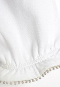 Tessuto bianco con una superficie testurizzata, caratterizzato da un elastico arricciato nella parte superiore e una rifinitura decorativa in argento con piccoli anelli lungo il bordo inferiore.