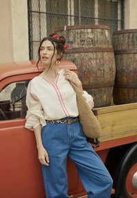 Donna con bandana rossa e blusa bianca si appoggia a un camion rosso accanto a barili di legno contrassegnati con il nome del ristorante.