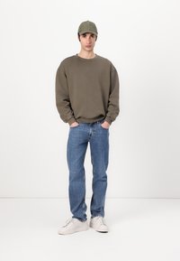 Sweat-shirt vert olive, jeans en denim bleu, baskets blanches et casquette verte. Coupe oversized avec des poignets côtelés et un style décontracté.
