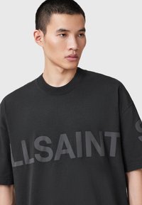 Tânăr cu părul scurt, purtând un tricou negru larg, cu textul mare "ALLSAINTS" parțial vizibil pe piept.