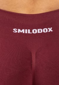 Zbliżenie na bordowe legginsy z prążkowaną teksturą i białym logo "SMILODOX" na pasku w talii.